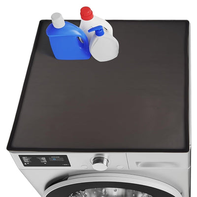 Black silicone washer and dryer top mat with raised edges, placed on a front load machine with laundry detergent bottles.
